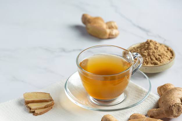 Glass cup of ginger tea served with fresh ginger slices and ginger powder on a light marble surface.