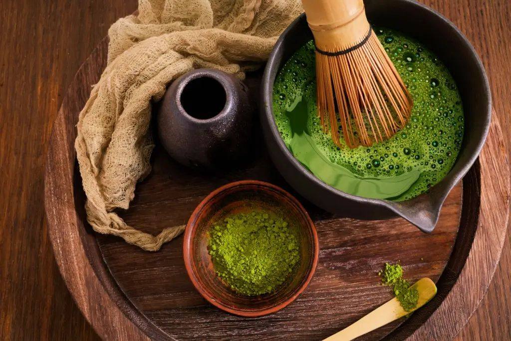 Japanese matcha tea preparation featuring a bamboo whisk, matcha bowl filled with frothy green tea, and powdered matcha on a wooden tray.