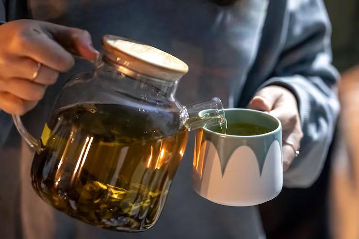 Person pouring freshly brewed herbal tea from a glass teapot into a ceramic cup, showcasing the tea brewing process and warm wellness ritual.